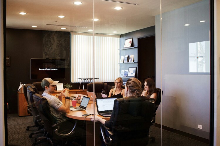 Group Of Professionals Sitting Around A Boardroom Table In A Glass Office With Laptops